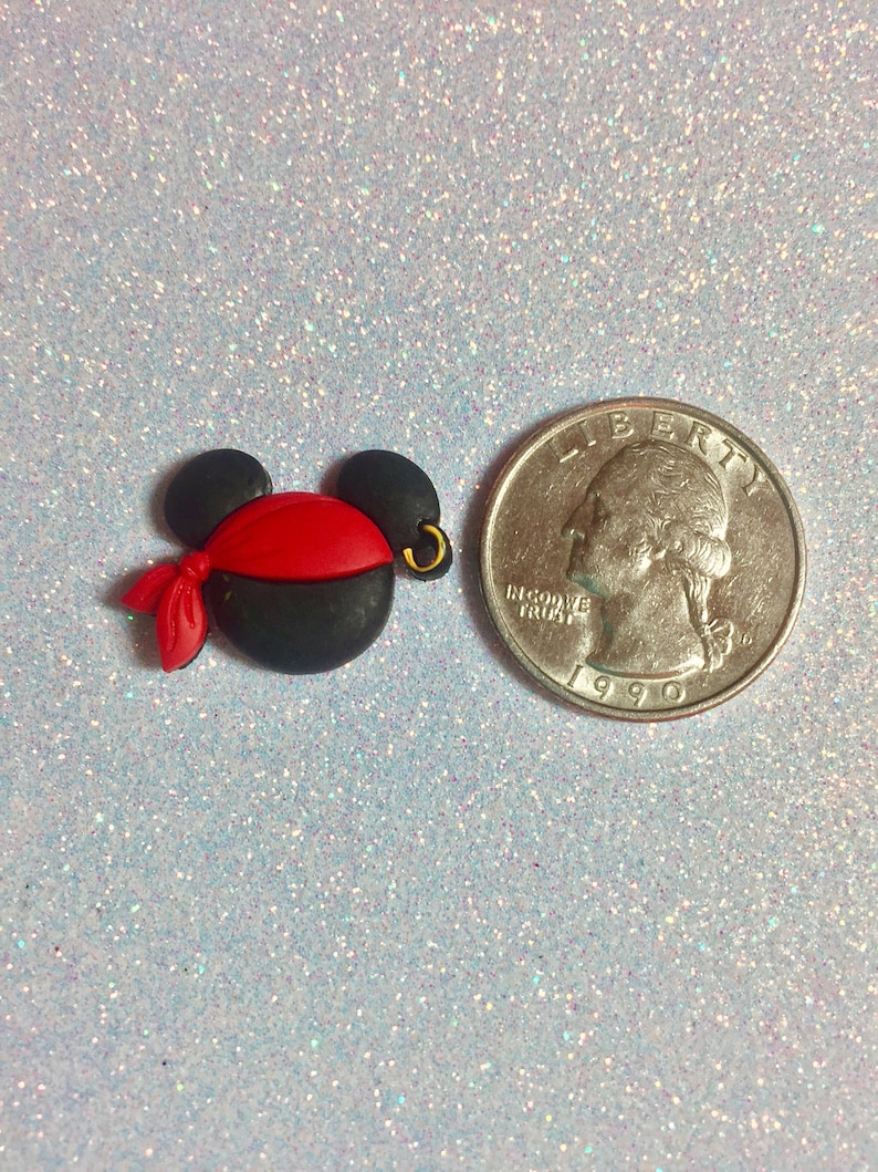May include: A black and red pirate-themed Mickey Mouse head charm with a gold hoop earring. The charm is next to a U.S. quarter coin for size comparison. The charm has a red bandana and is made of a smooth material.