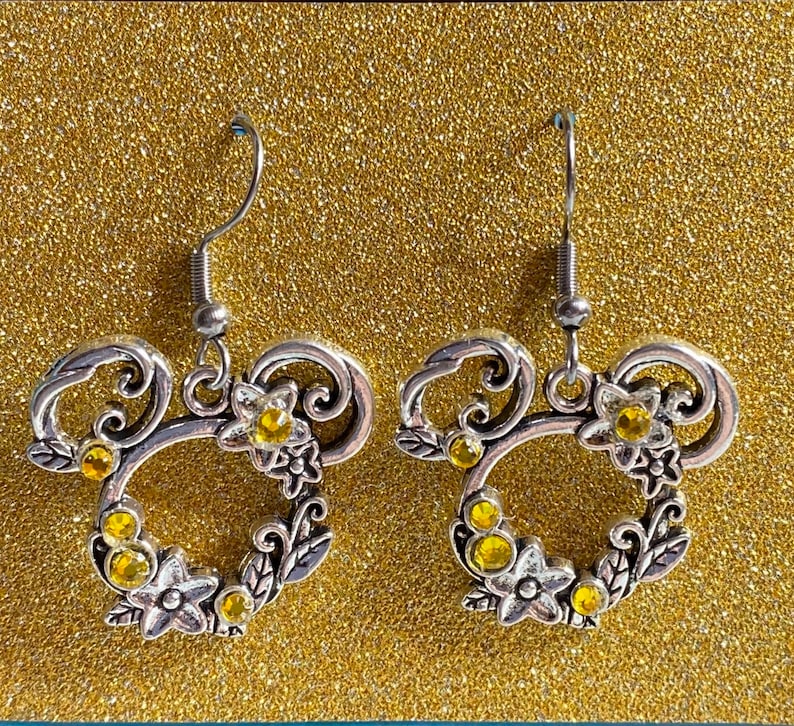 May include: A pair of silver earrings featuring a Mickey Mouse head design. The earrings are adorned with yellow rhinestones and have a floral pattern.