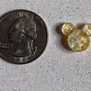 Op de afbeelding: Een klein, geel, iriserend, hars Mickey Mouse hoofd met een stippenpatroon. Het Mickey Mouse hoofd is ongeveer zo groot als een Nederlandse 2 euro munt.