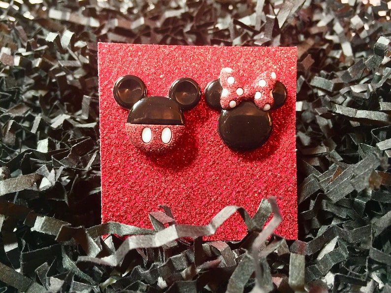 May include: A pair of red and black Mickey Mouse and Minnie Mouse character earrings. The earrings are on a red glitter background.