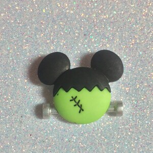 May include: Green and black Frankenstein Mickey Mouse hair clip. The clip has a green face with black stitches and black ears.