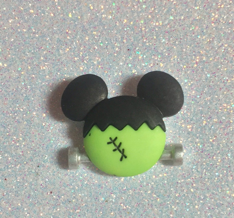 May include: Green and black Frankenstein Mickey Mouse hair clip. The clip has a green face with black stitches and black ears.