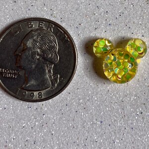 Op de afbeelding: Een gele Mickey Mouse-kopbedel met groene en gele glitter ingebed in hars. De bedel is ongeveer zo groot als een Nederlandse 2 euro munt.