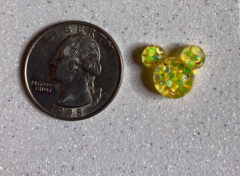 Op de afbeelding: Een gele Mickey Mouse-kopbedel met groene en gele glitter ingebed in hars. De bedel is ongeveer zo groot als een Nederlandse 2 euro munt.