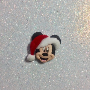 May include: A red and white Santa hat with a black Mickey Mouse face on a white glitter background.