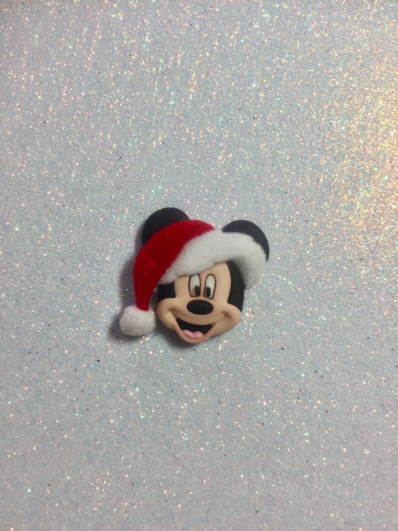 May include: A red and white Santa hat with a black Mickey Mouse face on a white glitter background.
