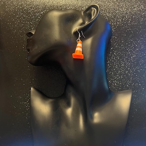 May include: A pair of novelty earrings featuring miniature orange and white striped traffic cones. The earrings are suspended from silver-colored fish hook ear wires. The cones have a bright orange base and white stripes.