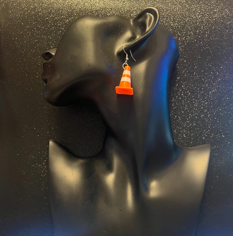 May include: A pair of novelty earrings featuring miniature orange and white striped traffic cones. The earrings are suspended from silver-colored fish hook ear wires. The cones have a bright orange base and white stripes.
