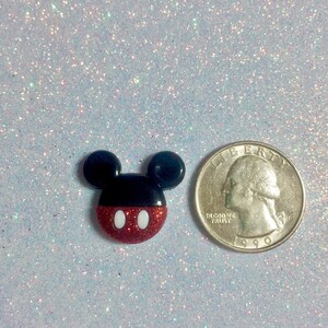 May include: A red, white, and black Mickey Mouse character charm with glitter. The charm is about the size of a US quarter.