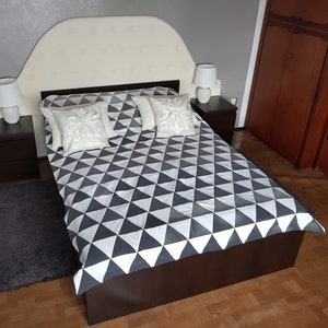 May include: A white headboard with a curved top sits above a dark brown bed frame with a white and gray geometric patterned duvet. Two white pillows with embroidered floral designs are on the bed. Two bedside tables with lamps are on either side of the bed.