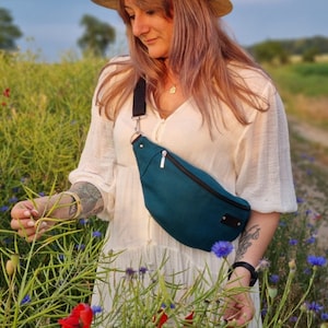 May include: A woman wearing a white dress and a straw hat is standing in a field of wildflowers. She is holding a teal blue fanny pack with a black strap.