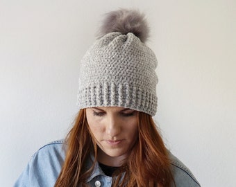 May include: A grey crocheted beanie with a fluffy pom pom. The beanie is being worn by a person with long red hair and a denim jacket.