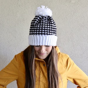 May include: A black and white houndstooth crochet beanie with a white pom pom on top. The beanie is worn by a person wearing a mustard yellow jacket.