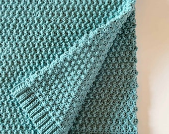 May include: A teal-colored crocheted blanket featuring a textured bobble stitch pattern throughout the main body. The blanket is finished with a ribbed border along the edges, showcasing a consistent yarn color and uniform stitch construction.