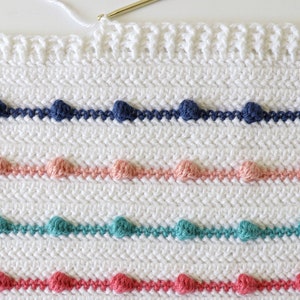 Puede incluir: Una manta blanca de ganchillo con filas de detalles de punto de pomp&oacute;n de colores. La manta presenta filas de pompones azules, rosas, verde azulado y rojos.