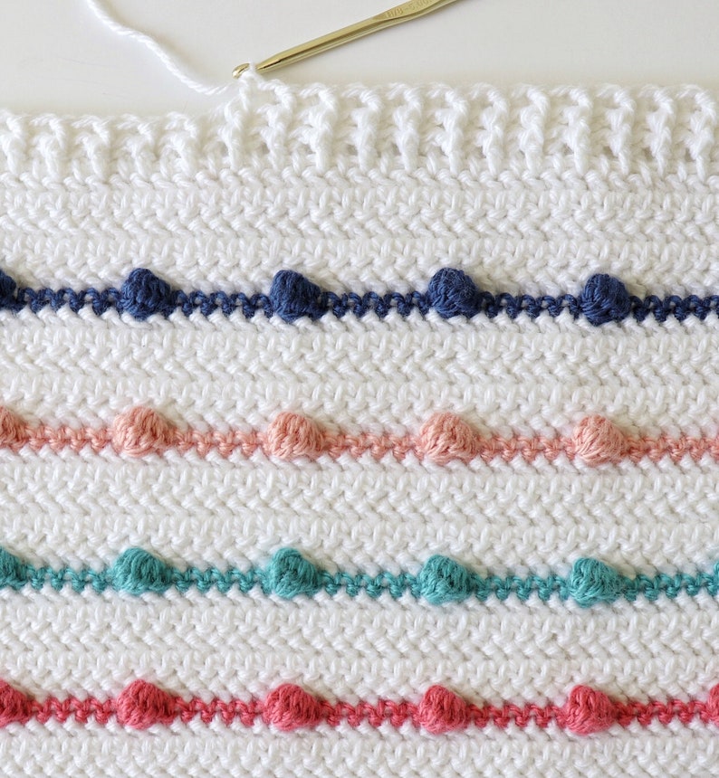 Puede incluir: Una manta blanca de ganchillo con filas de detalles de punto de pomp&oacute;n de colores. La manta presenta filas de pompones azules, rosas, verde azulado y rojos.
