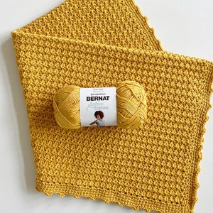 Pu&ograve; includere: Una copertina per neonati in crochet giallo con un gomitolo di lana giallo e l'etichetta "Bernat Softee Cotton" su di essa.