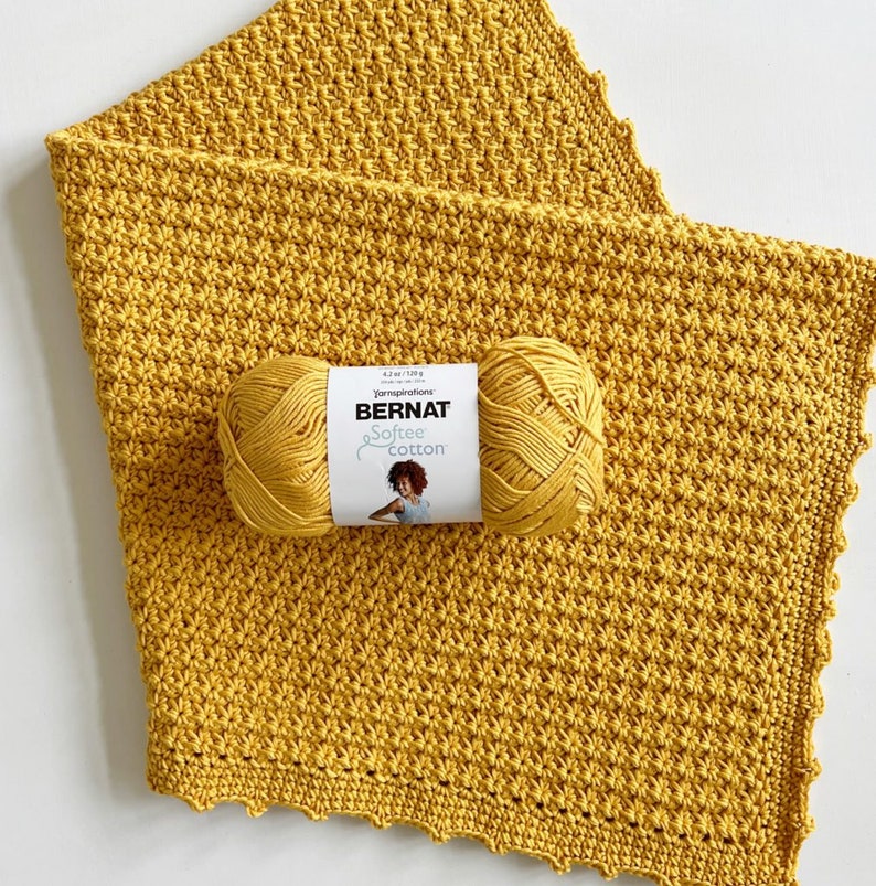 Pu&ograve; includere: Una copertina per neonati in crochet giallo con un gomitolo di lana giallo e l'etichetta "Bernat Softee Cotton" su di essa.
