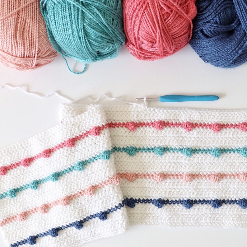 Puede incluir: Una manta de crochet blanca con filas de hilo rosa, turquesa y azul marino. La manta est&aacute; hecha con un punto de pomp&oacute;n y tiene un dise&ntilde;o simple y moderno.