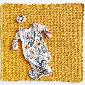 Pu&ograve; includere: Una copertina per neonati in crochet color giallo senape con un vestito annodato a stampa floreale e un fiocco per capelli in cima.
