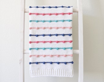 Può includere: Una copertina per neonati in crochet bianco con strisce colorate e pompon. La copertina è appesa a una scala bianca.