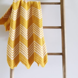 Può includere: Una copertina per neonati in crochet gialla e bianca a chevron drappeggiata su una scala di legno.