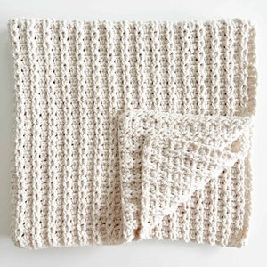 May include: Cream-colored, hand-crocheted blanket with a textured, ribbed pattern. The blanket is folded, revealing a smaller, matching square. The soft, neutral tones suggest a cozy, handmade item, perfect for home decor or baby items.