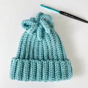 May include: A teal blue crocheted beanie with a knotted top. The beanie is lying on a white surface.
