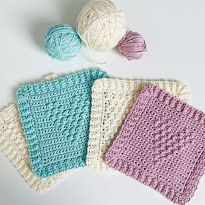 May include: Four crocheted dishcloths in different colors: light blue, cream, and light purple. The dishcloths are arranged in a row with the cream dishcloth in the middle. There are three balls of yarn in the background: cream, light blue, and light purple.