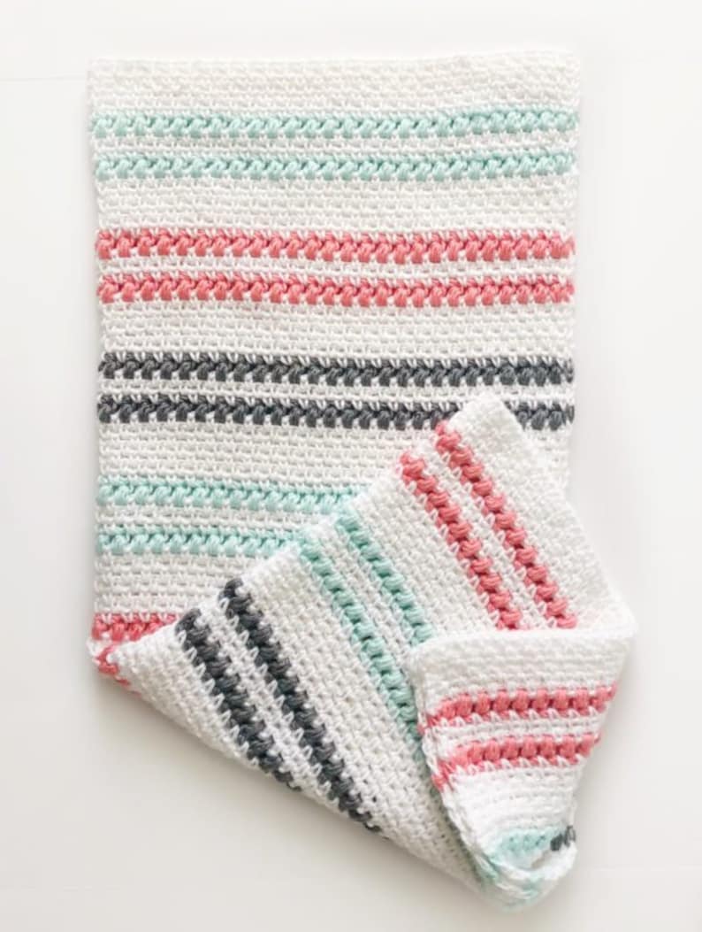 May include: A white crocheted blanket with stripes of light blue, pink, and gray yarn. The blanket is folded in half and the edges are visible.