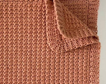 May include: A crocheted blanket in a warm terracotta color. The blanket is folded in half, with one corner folded over. The blanket is made with a simple stitch pattern, creating a textured look.
