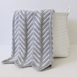 Puede incluir: Una manta de crochet con patrón de chevron gris y blanco doblada sobre el borde de una cesta tejida blanca.