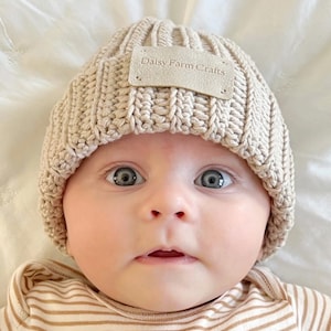 May include: A baby wearing a beige knitted beanie with a brown leather patch that says "Daisy Farm Crafts".