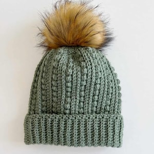 May include: A sage green knit beanie with a ribbed brim and a textured stitch pattern. The hat is topped with a large, fluffy faux fur pom-pom in shades of brown and black. This winter hat is a cozy accessory.