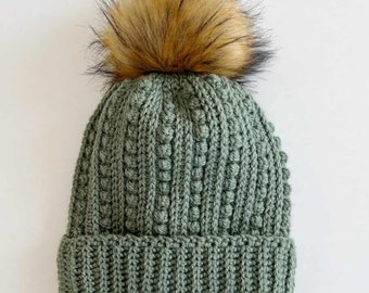 May include: A sage green knitted beanie with a ribbed brim and a textured stitch pattern. The hat features a large, fluffy faux fur pom-pom in brown and black. This winter hat is a warm accessory, perfect for cold weather.
