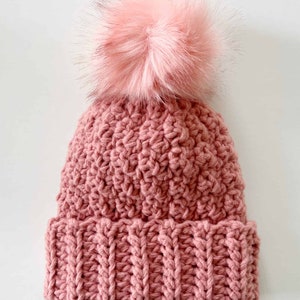 Puede incluir: Un gorro de punto rosa con un pompón rosa esponjoso en la parte superior.