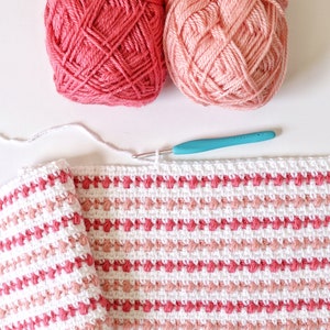 Könnte beinhalten: Nahaufnahme einer gehäkelten Decke mit rosa und weißen Streifen. Die Decke wird mit einer Häkelnadel und weißem Garn gearbeitet.