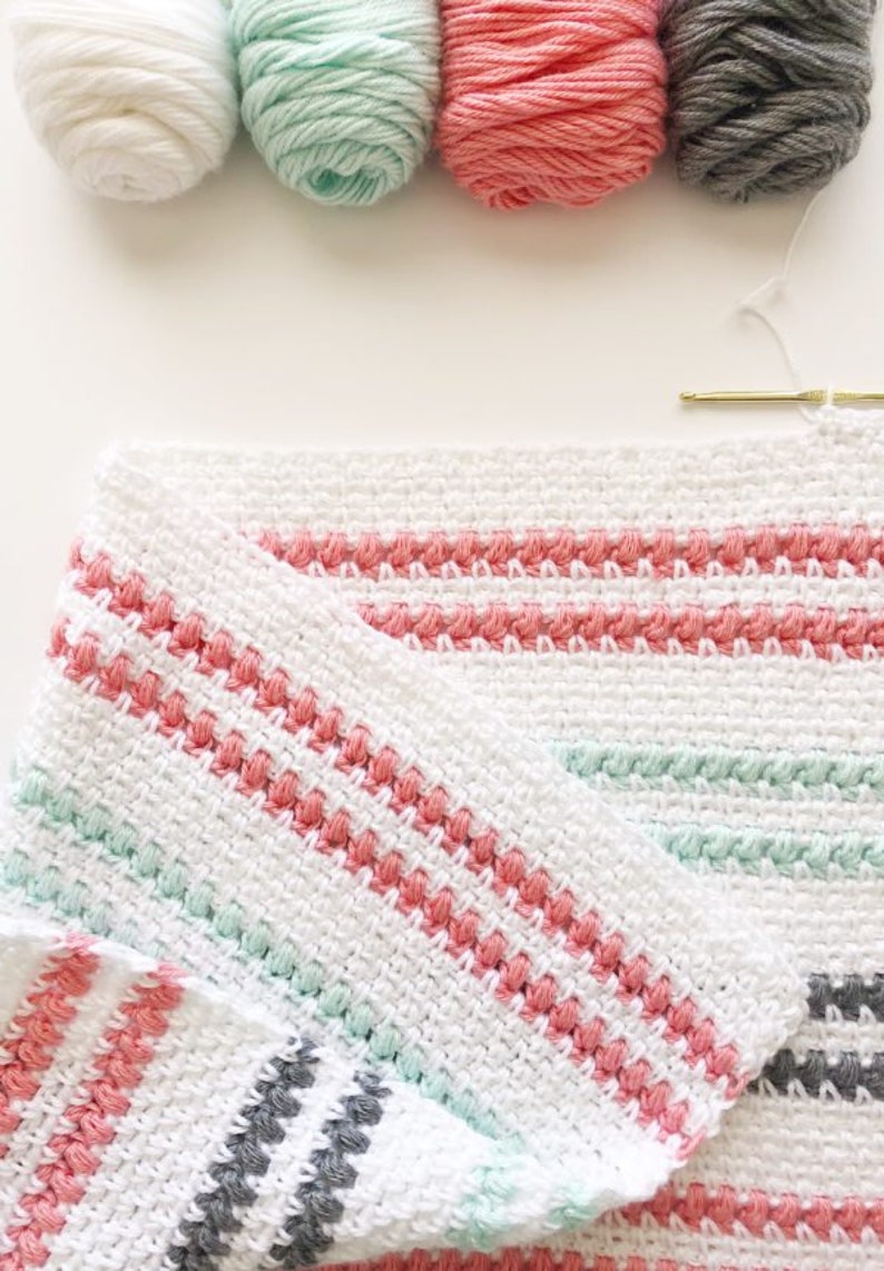 May include: A white crochet blanket with stripes of pink, mint green, and gray yarn. The blanket is folded in half and the yarn balls used to make the blanket are visible in the top of the image.
