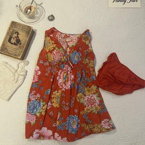 Könnte beinhalten: Ein rotes Kleid mit Blumenmuster und passendem Unterteil. Das Kleid hat einen V-Ausschnitt und einen Kordelzug, verziert mit rosa, blauen und gelben Blumen. Ein Vintage-Buch, eine Teetasse und Schmuck sind ebenfalls vorhanden.