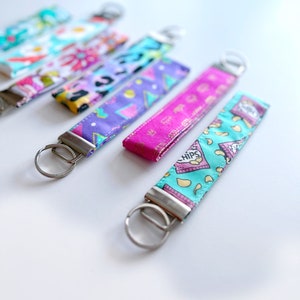 May include: A set of six colourful keychains with different patterns. The keychains are made of fabric and have silver key rings. The patterns include a pink and purple geometric design, a blue and white floral design, a pink and white floral design, a blue and white chip bag design, and a green and white chip bag design.