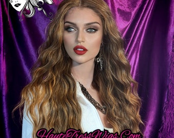 May include: A long, wavy, light brown human hair wig with a slight blonde highlight. The wig is styled in loose waves and falls to the shoulders. The model is wearing a white top and has a natural makeup look with red lipstick.