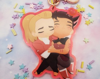 May include: A pink and white acrylic keychain featuring a cartoon couple hugging. The characters are a woman with blonde hair and a man with brown hair and cat ears. The keychain has a gold heart-shaped clasp.