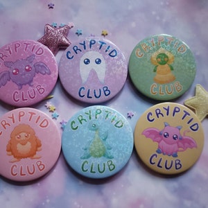 Op de afbeelding: Zes ronde, kleurrijke knoppen met de tekst "Cryptid Club" en illustraties van verschillende cryptiden. De knoppen zijn roze, paars, groen, geel, blauw en oranje.