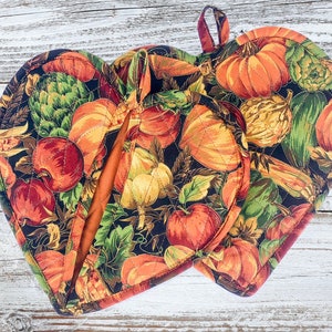 May include: Heart-shaped oven gloves with a fall harvest pattern featuring pumpkins, apples, and gourds. The gloves are on a white wooden surface, with decorative corn, pinecones, and acorns in the background.
