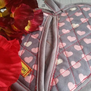 Puede incluir: Un guante de horno gris con un patrón de corazones rosas y blancos. El guante tiene un bolsillo en la parte posterior y está acolchado con costuras rojas.