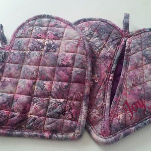 Puede incluir: Un par de agarraderas de horno con forma de corazón acolchadas en un patrón floral rosa y morado. Las agarraderas tienen un lazo para colgar y una tiene la palabra "Alegría" bordada.