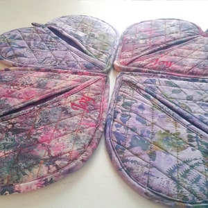 Op de afbeelding: Vier hartvormige ovenwanten in paars en roze met een gestikt ontwerp. Elke ovenwant heeft een zakje en is geborduurd met het woord "Vreugde".