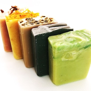 May include: Five bars of handmade soap in a row, from left to right: pink, yellow, beige, dark gray, and light green. The soap bars are arranged on a white surface.