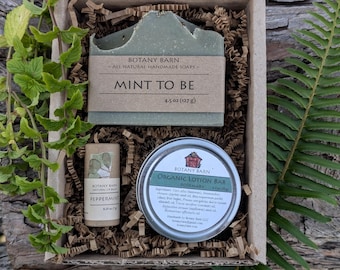 May include: A gift box containing a rectangular bar of green soap labeled Botany Barn Mint To Be 4.5 oz, a cylindrical peppermint lip balm, and a round tin of organic rosemary lotion bar, all nestled in brown paper crinkle filler.