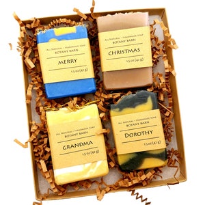 May include: A gift box filled with brown paper shreds contains four bars of handmade soap. The soap bars are labeled "Merry", "Christmas", "Grandma", and "Dorothy". Each bar is 1.5 ounces (42 grams).